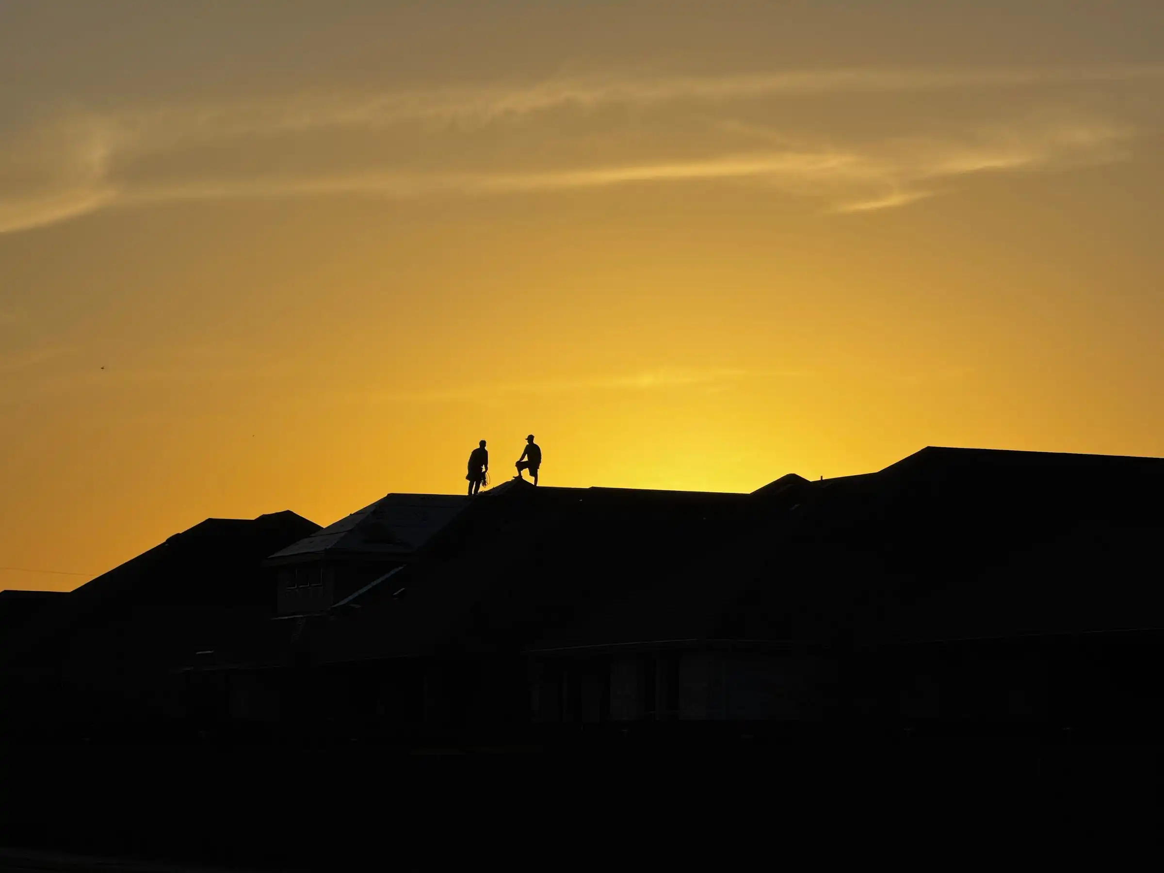 Roofers silhouetted against a sunset, working along the ridge of a roof.